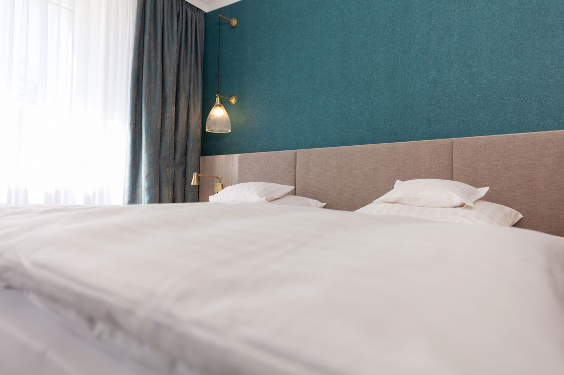 A neatly made bed with white bed linen and pillows in a modern room with a teal-coloured accent wall, a beige headboard and a golden wall lamp next to a window with white curtains and dark curtains.