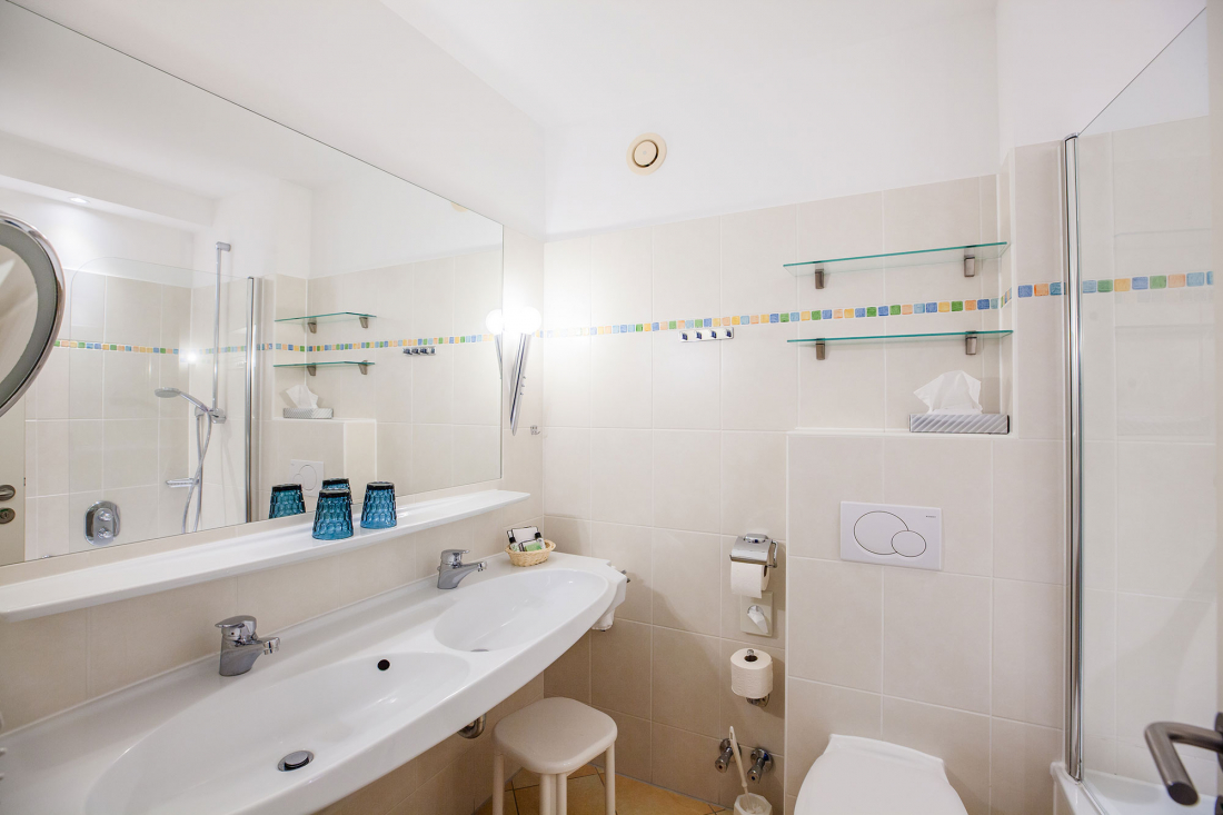 Helles, modernes Badezimmer mit Doppelwaschbecken, großem Spiegel, Glasablagen, Tissue-Box und Toilettenartikeln. Beigefarbene Fliesen und bunte Verzierungen betonen den sauberen, minimalistischen Raum. Dusche und Toilette sind ebenfalls sichtbar.