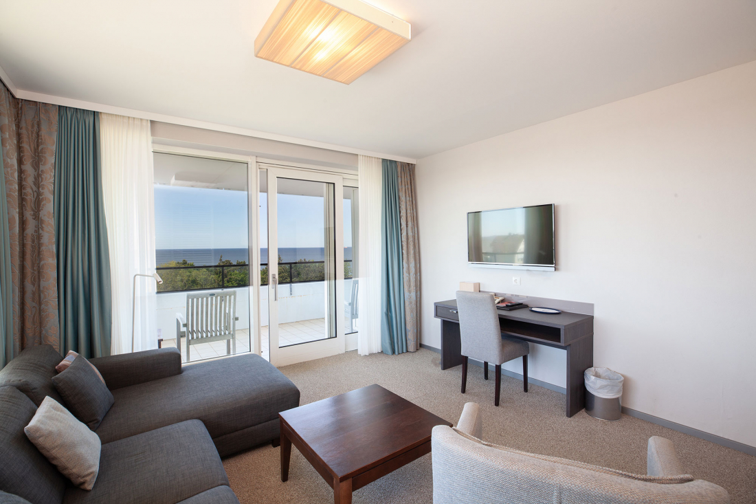 Helles Wohnzimmer der Zwei-Raum-Suite mit einem Sofa, einem Couchtisch, einem Schreibtisch und einem an der Wand montierten Fernseher. Große Glastüren führen auf einen Balkon mit Stühlen und bieten einen Blick auf das Meer und das Grün draußen.