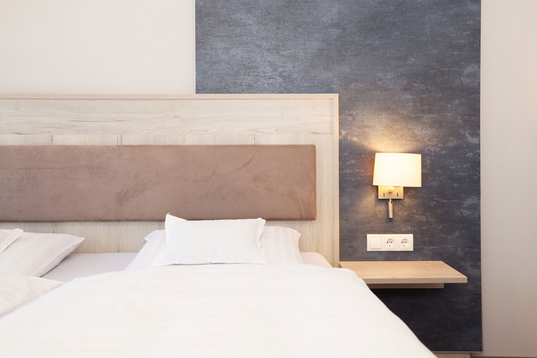 Ein ordentlich gemachtes Bett mit weißer Bettwäsche und einem hellbraun gepolsterten Kopfteil steht neben einer Wandlampe über einem kleinen Holzregal vor einer blauen Akzentwand. Zwei Steckdosen befinden sich unter der Lampe.