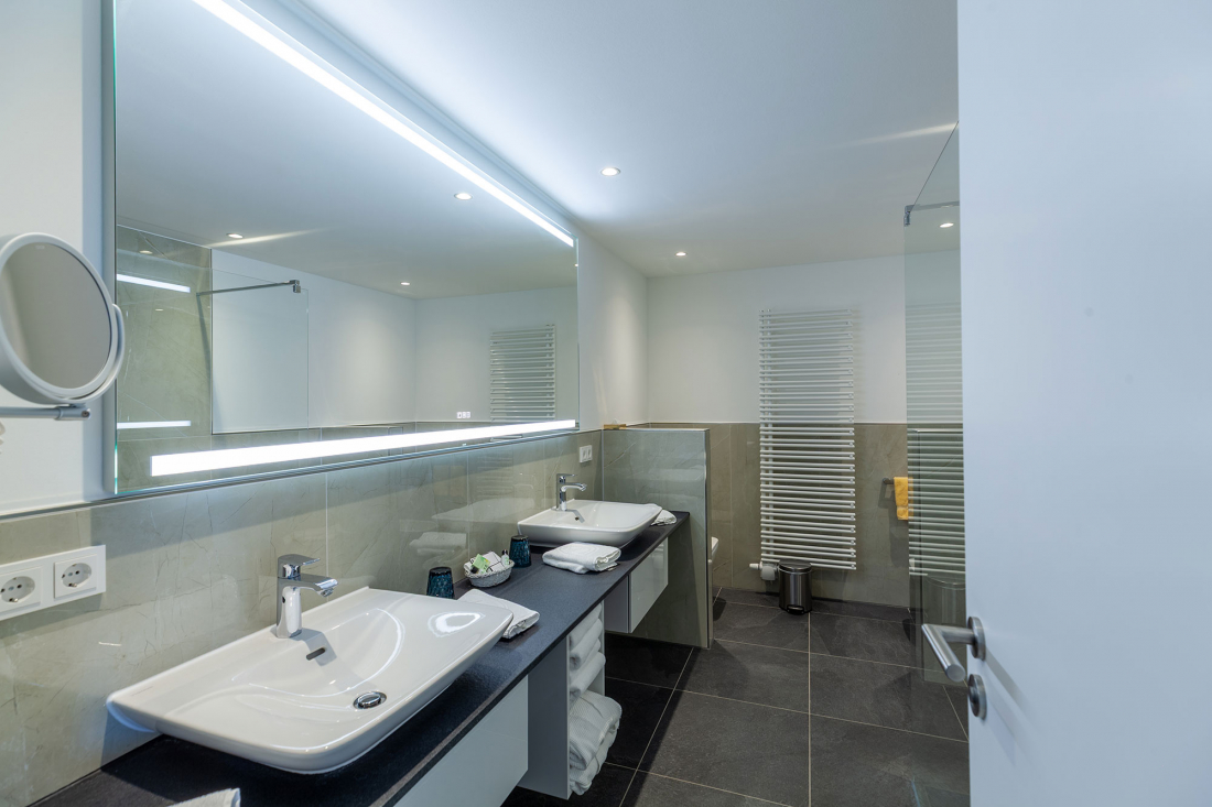 Modernes Badezimmer mit einem großen Spiegel über zwei Waschbecken, schwarzer Arbeitsplatte, weißen Handtüchern und Toilettenartikeln. Der Raum verfügt über graue Bodenfliesen, einen beheizten Handtuchhalter und eine teilweise offene Tür.