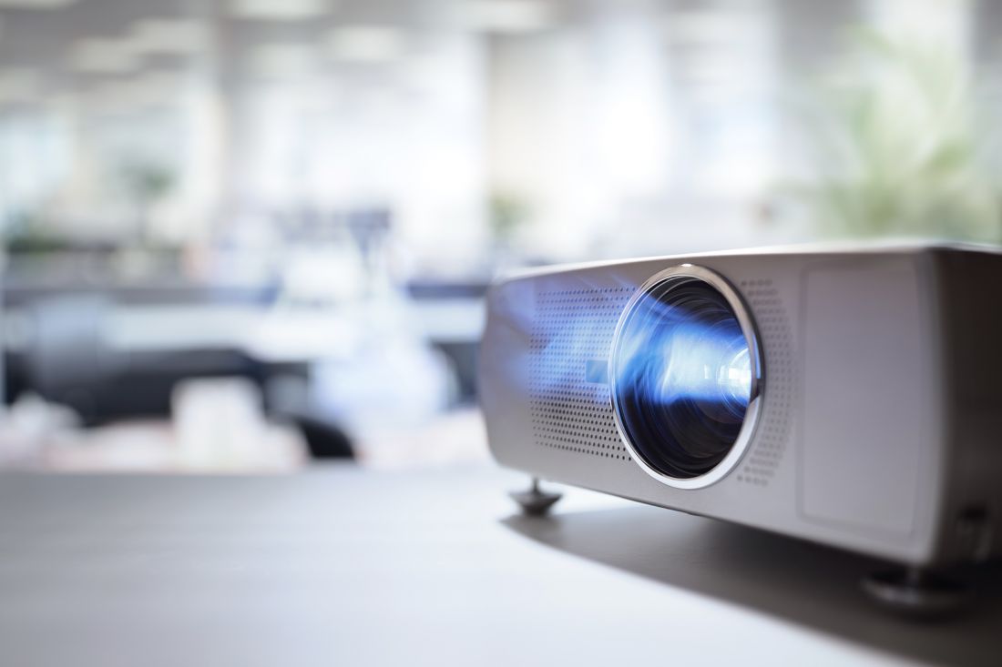 Close-up of a digital projector on a table in a bright, modern office, with its lens emitting blue light and a blurred background of desks and lights.