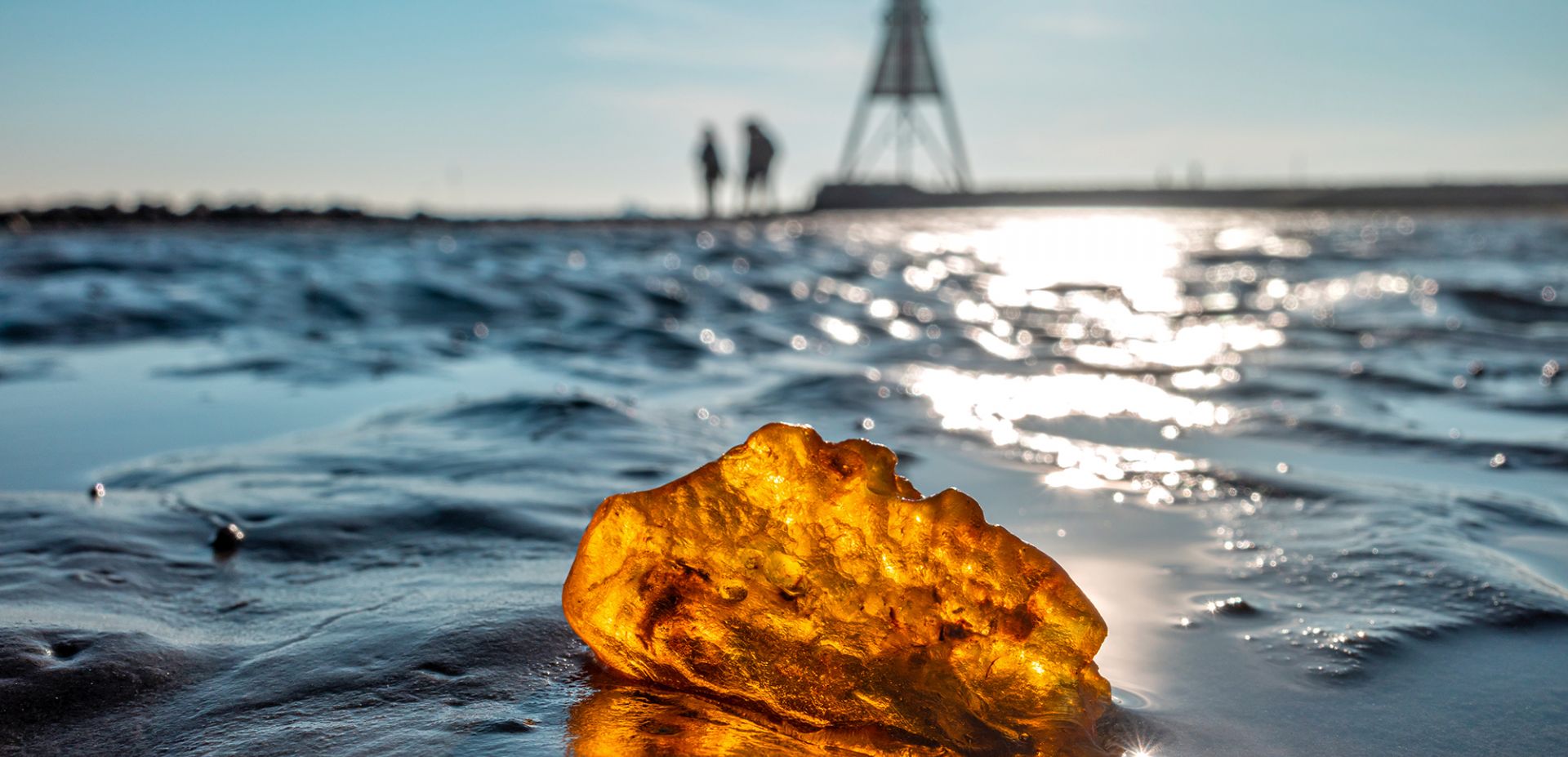 Ein großes Stück Bernstein liegt im nassen Watt an der Küste und glitzert im Sonnenlicht. Im Hintergrund gehen zwei unscharfe Gestalten unter strahlend blauem Himmel auf die Kugelbake zu.
