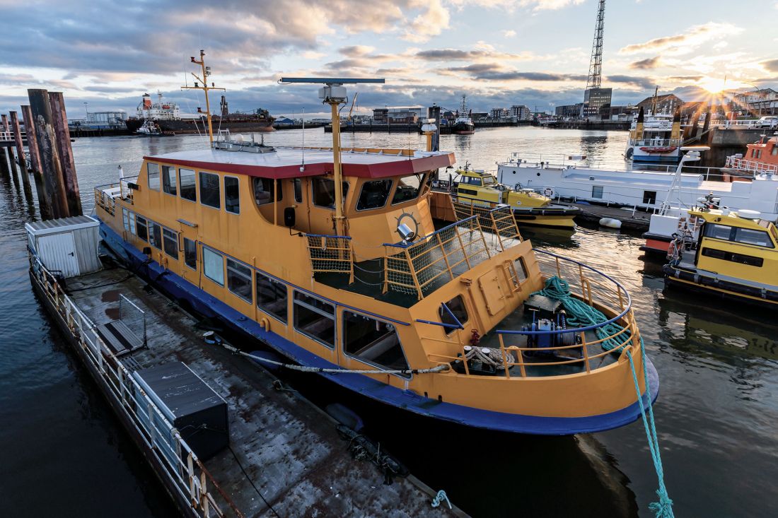 Eine gelb-blaue Fähre liegt im Hafen, umgeben von anderen Booten. Die Sonne geht im Hintergrund unter und wirft ein warmes Licht auf das Wasser und die Gebäude.