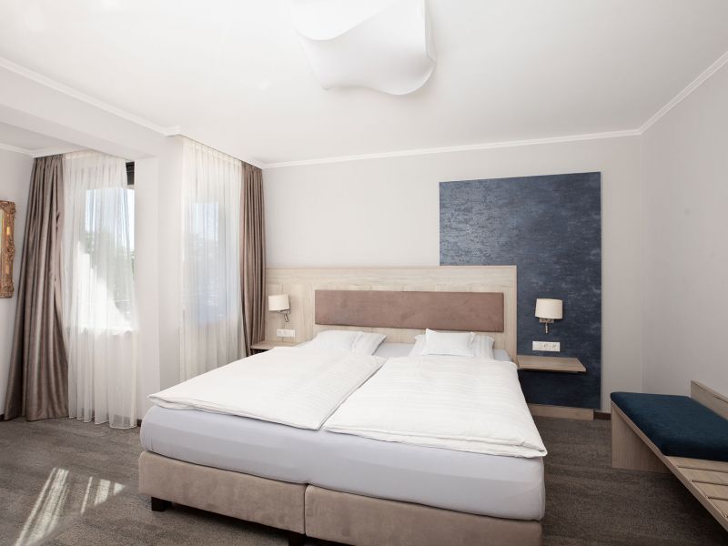 A modern hotel room in the three-room flat with a large double bed, white bed linen, two bedside tables with lamps, a blue accent wall, large windows with curtains and a bench against one wall.