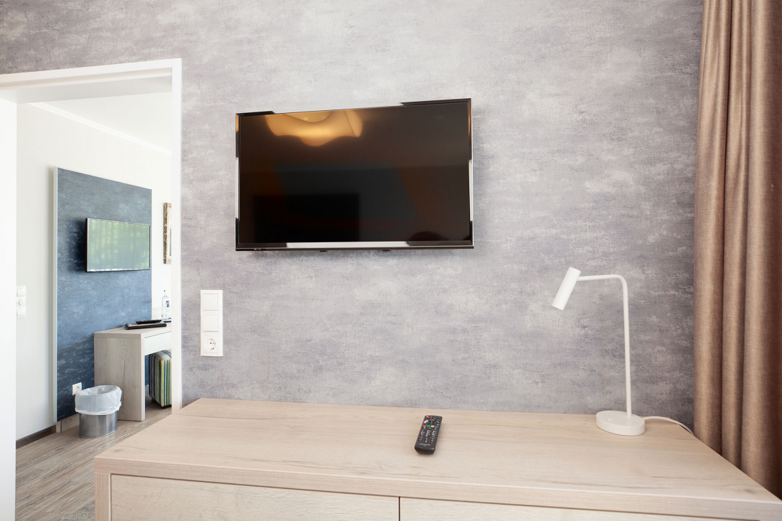 Ein Flachbildfernseher ist an einer strukturierten grauen Wand über einer hellen Holzkommode mit einer Fernbedienung und einer weißen Schreibtischlampe angebracht. Ein weiterer Fernseher und ein Mülleimer sind im angrenzenden Raum zu sehen.