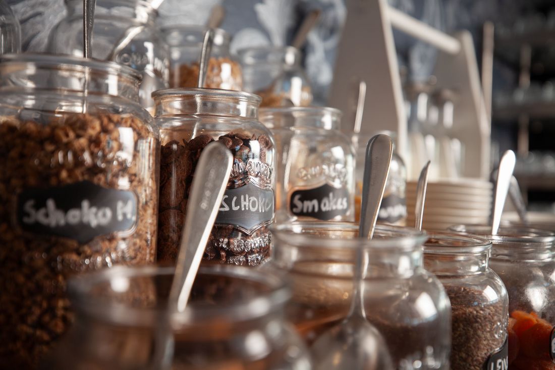 A series of jars with different cereals and toppings, each with a spoon and a small chalkboard label, placed on a breakfast bar or buffet.