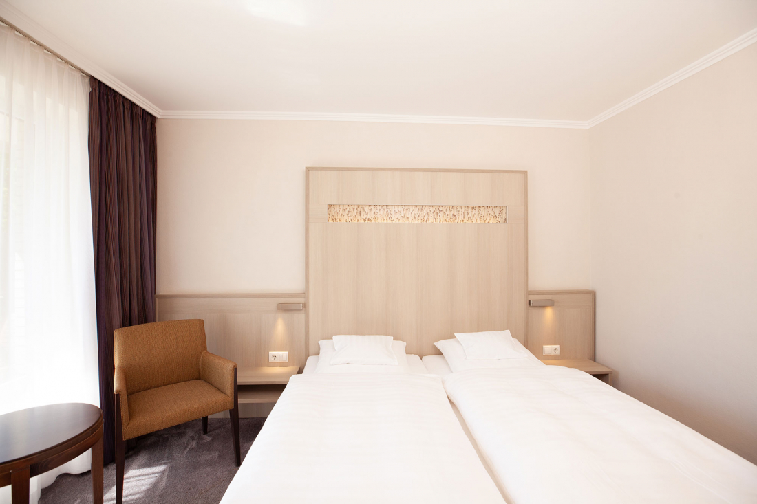A bright, modern hotel room with a double bed, white bedding, a wooden headboard, a brown armchair, a small round table and a large window with transparent and dark curtains.