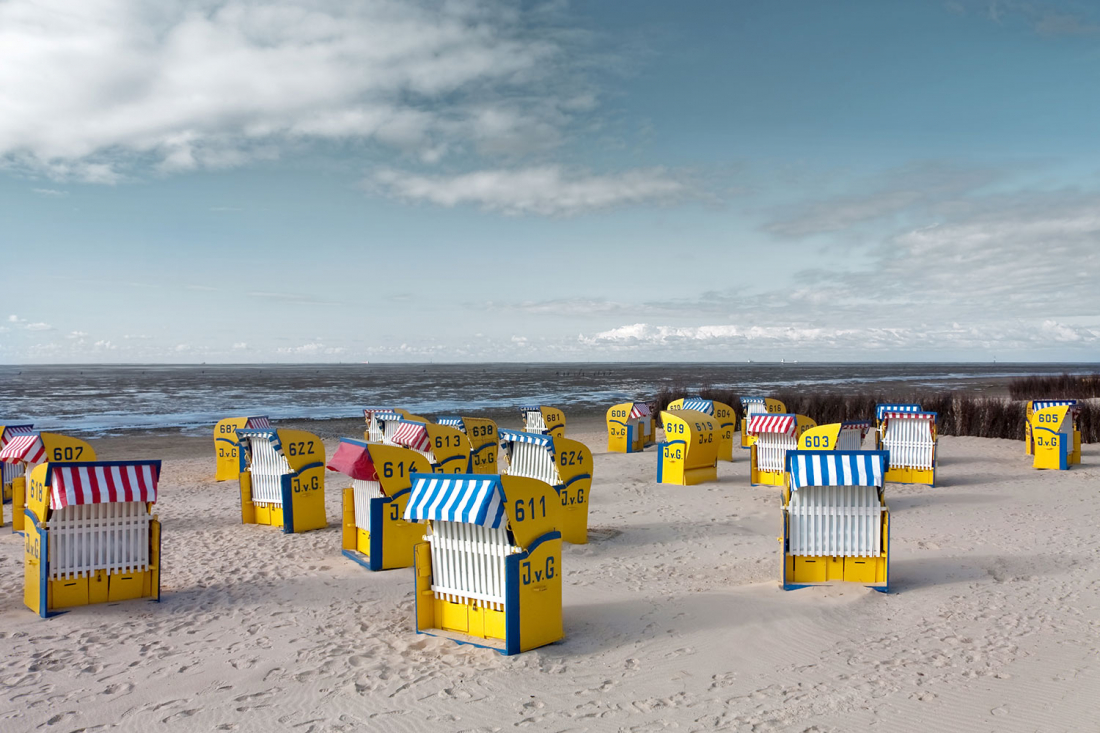 Gelbe Strandkörbe mit gestreiften blauen oder roten Schirmen sind an einem Sandstrand mit Blick auf das Meer unter einem teilweise bewölkten Himmel angeordnet. Jeder Stuhl trägt eine Nummer und die Initialen J.v.G.