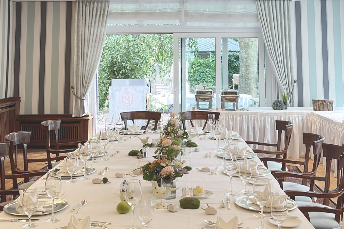 Meeting Room 1, "Garden Breeze," features striped wallpaper, large windows, wooden chairs, and a long table set with white linens, glasses, and napkins, ready for a formal meal. The greenery outside can be seen through the windows.