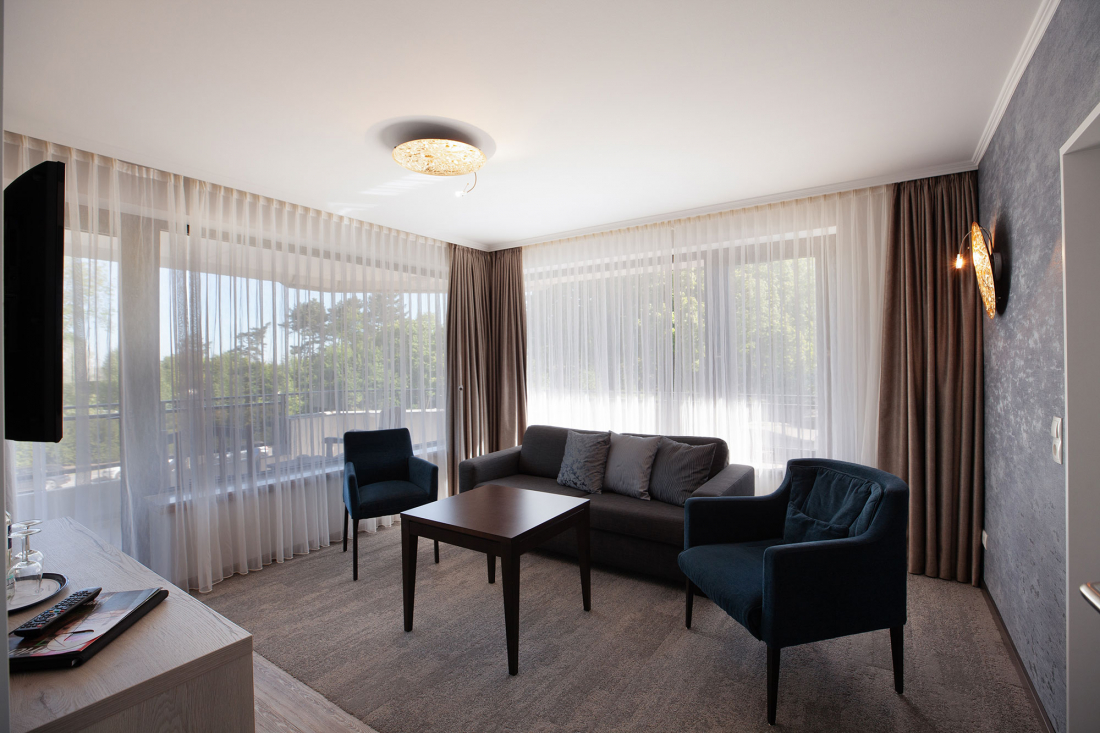 Ein helles Wohnzimmer eines Dreiraum-Appartement mit durchsichtigen Vorhängen, grauem Sofa mit Kissen, zwei dunkelblauen Sesseln und einem kleinen Couchtisch aus Holz. Große Fenster lassen natürliches Licht herein.