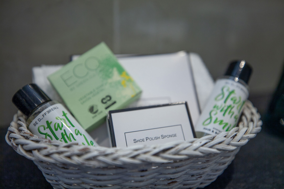 Ein weißer Weidenkorb enthält Toilettenartikel, darunter Flaschen mit der Aufschrift "Start the Day with a Smile" (Starte den Tag mit einem Lächeln), eine grüne ECO-Seifendose, eine Schuhputzschwammdose und gefaltete weiße Handtücher.