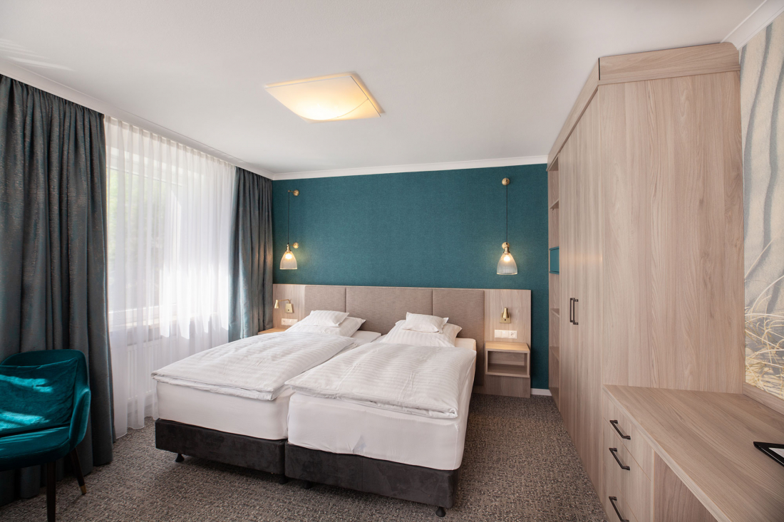 A modern hotel room with two single beds pushed together, teal-coloured accent wall, light wooden furniture, teal-coloured chair, large wardrobe and a window with blackout curtains that let in natural light.