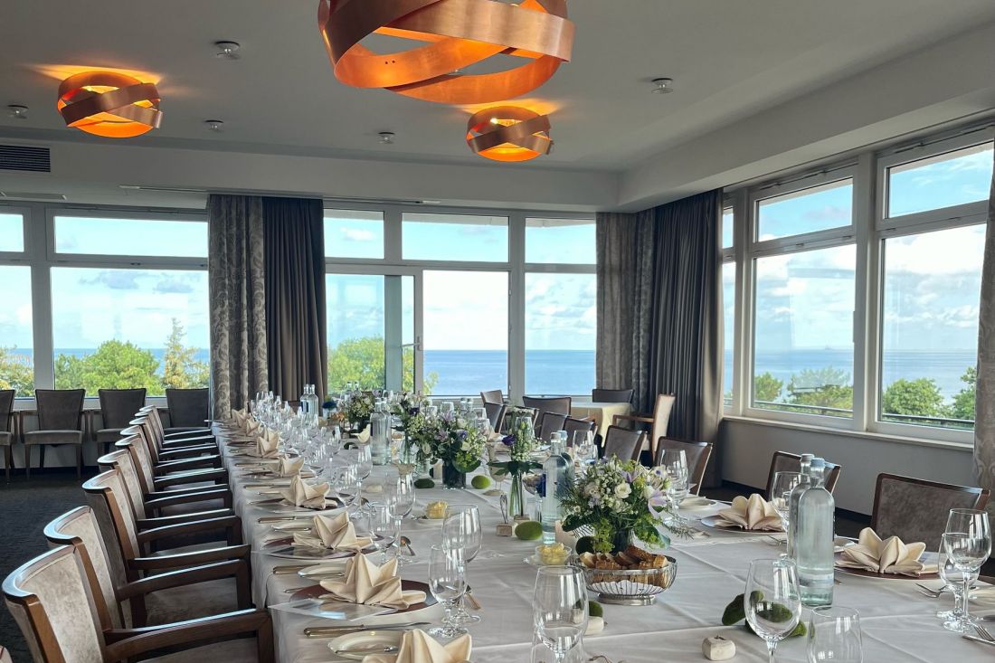 A long table set for an elegant event with white tablecloths, folded napkins, glasses, floral decorations and large windows offering a picturesque view of trees and the North Sea under a cloudless sky.