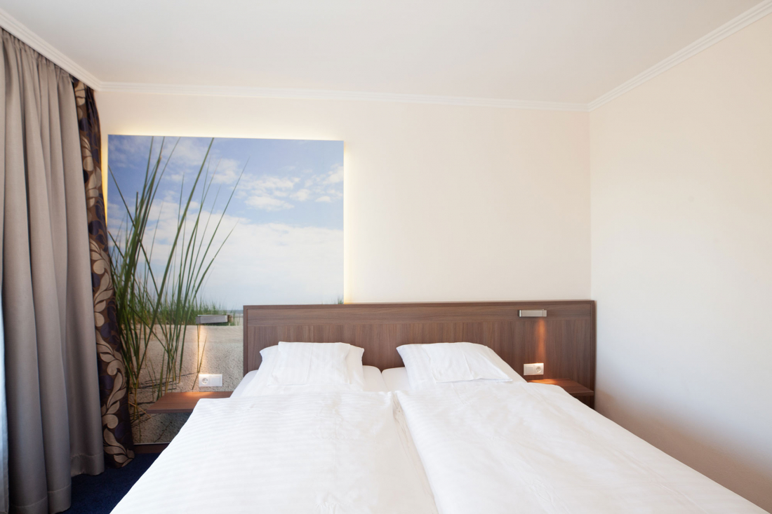 Ein modernes Zweiraum-Appartement mit zwei sauber gemachten Betten, einem hölzernen Kopfteil, hellen Wänden, einem großen Fenster mit Vorhängen und einem Naturmotiv über dem Bett.