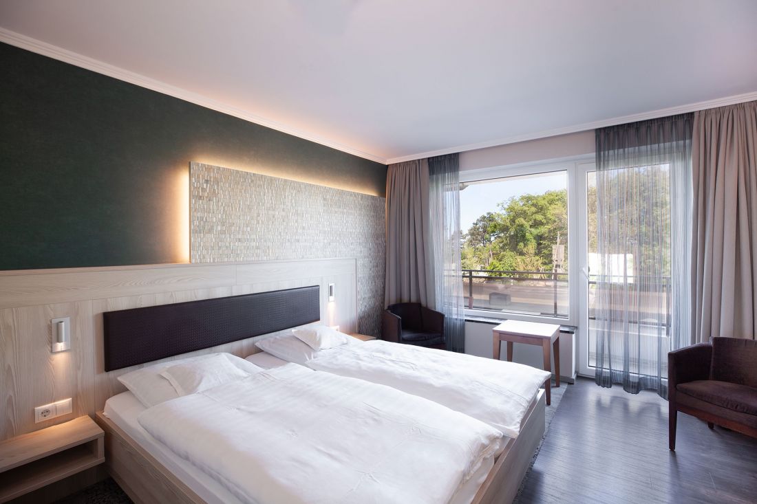 A modern double room with a double bed, white bedding, a wooden headboard with built-in lamps, a dark green accent wall, a large window with blackout curtains, chairs and a small table.