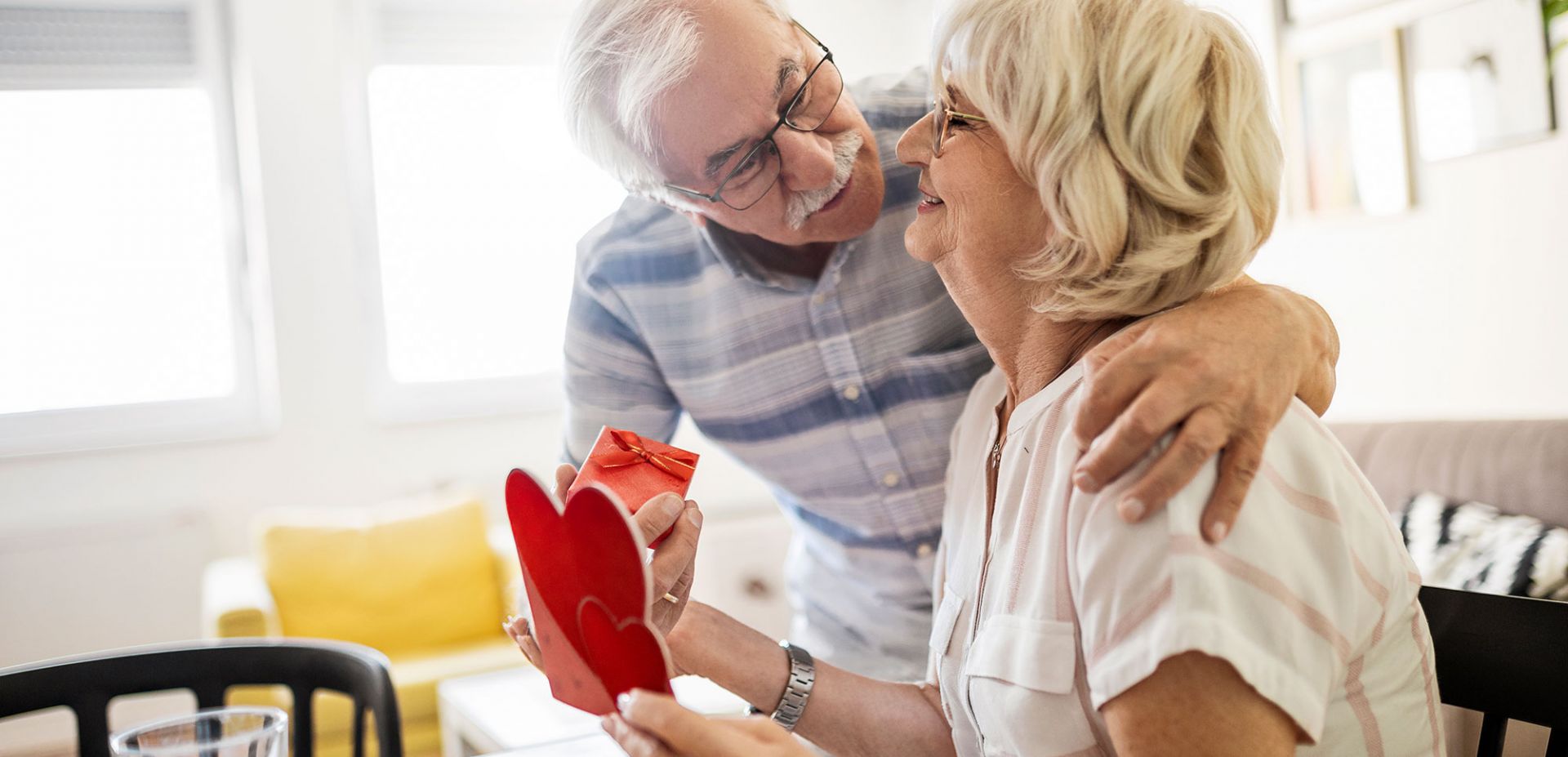 An older man hugs an older woman, who smiles and holds a red heart-shaped card in her hand. They sit at a table with coffee and juice in a bright, cosy living room and share a warm, tender moment.