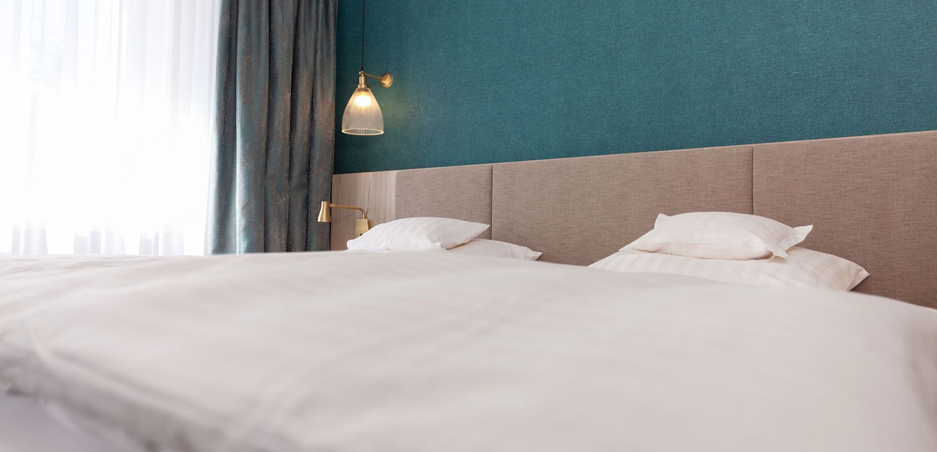 A neatly made bed with white bed linen and pillows in a modern room with a teal-coloured accent wall, a beige headboard and a golden wall lamp next to a window with white curtains and dark curtains.