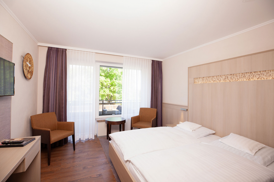 A bright hotel room with a double bed, two brown chairs, a small round table by a window with sheer curtains, a TV on the wall and light-coloured wooden furniture. There are green trees outside.