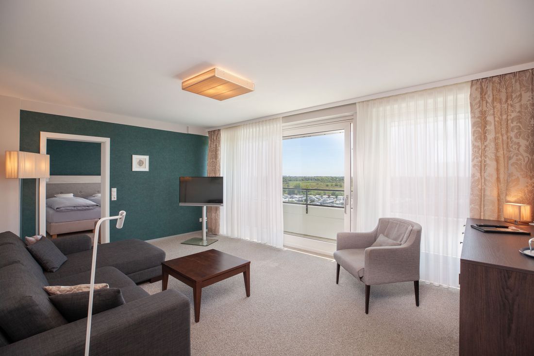 Wohnbereich der Drei-Raum-Suite mit einem grauen Sofa, einem Sessel, einem Couchtisch, einem Fernseher und einem großen Fenster mit durchsichtigen Vorhängen. Durch eine offene Tür ist ein Schlafzimmer mit einem Bett zu sehen.