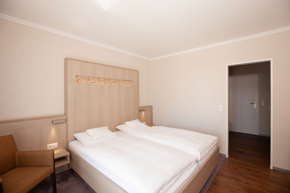 A minimalist double room with two single mattresses in a double bed, white bedding, a beige headboard, a brown armchair, wooden floor and neutral walls. An open door leads into the small entrance hallway.