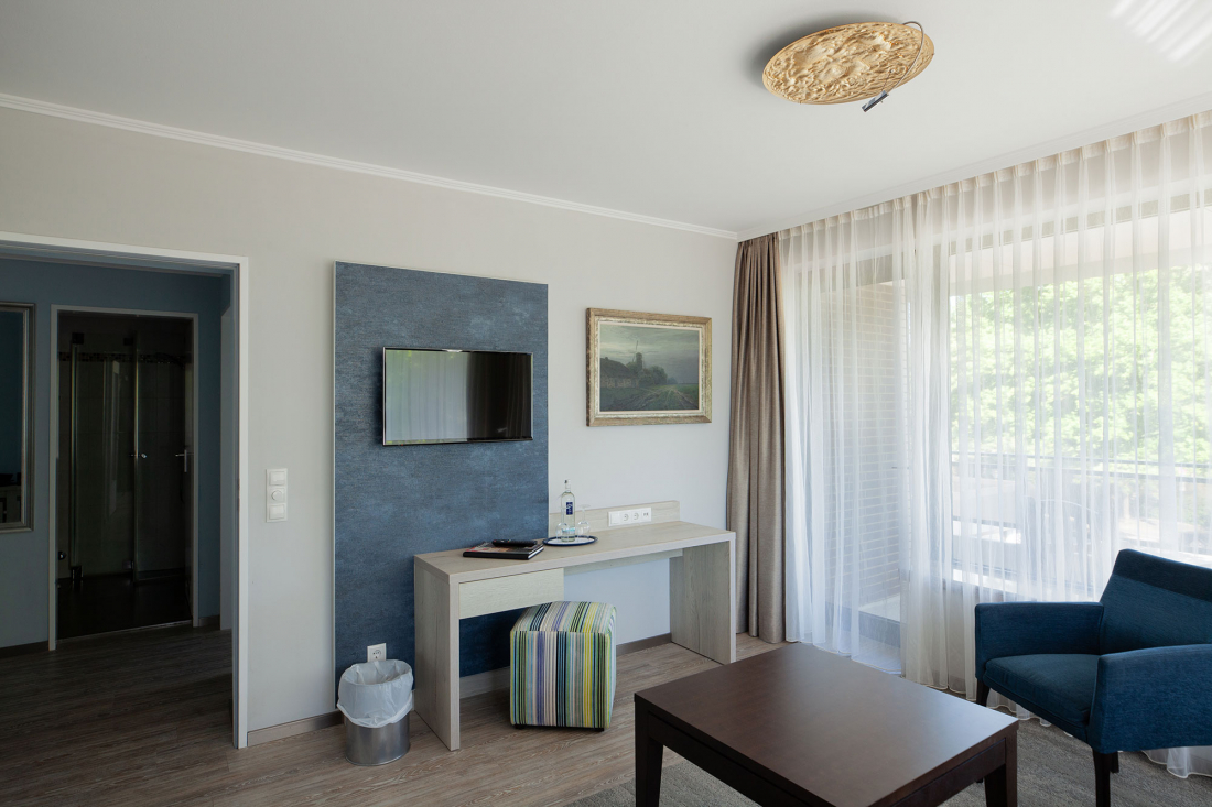 Ein modernes, lichtdurchflutetes Hotelzimmer mit einem eingebauten Fernseher, einem Schreibtisch, einem gestreiften Hocker, einem blauen Sessel, einem Couchtisch, großen Fenstern mit durchsichtigen Vorhängen und einer Tür, die zu einem Badezimmer führt.