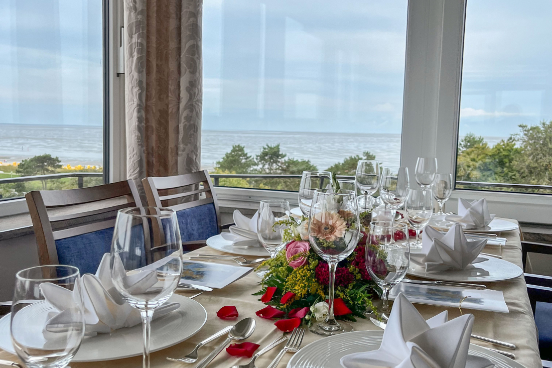 "Café zur Schönen Aussicht" A dining table set for a formal meal with folded napkins, glasses, and floral arrangements, placed by a large window overlooking trees and the sea. Red rose petals are scattered on the table.