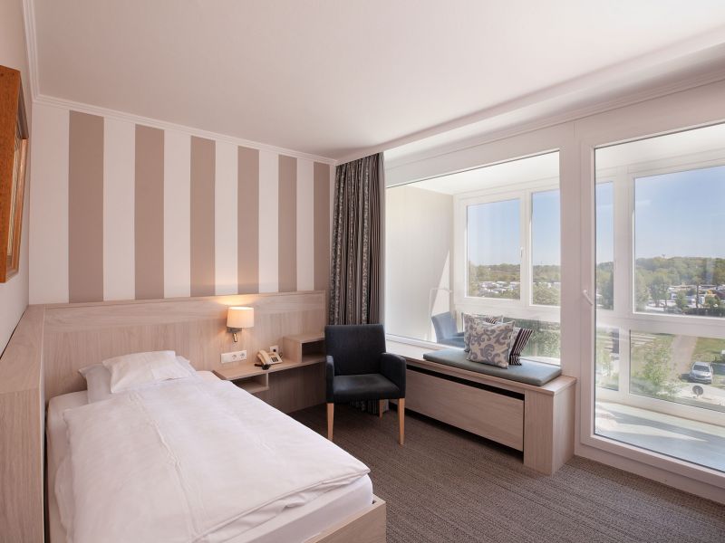A bright, modern hotel room with a single bed, a striped accent wall, a bedside table, an armchair and a large window seat overlooking a picturesque outdoor area. The room is flooded with natural light.