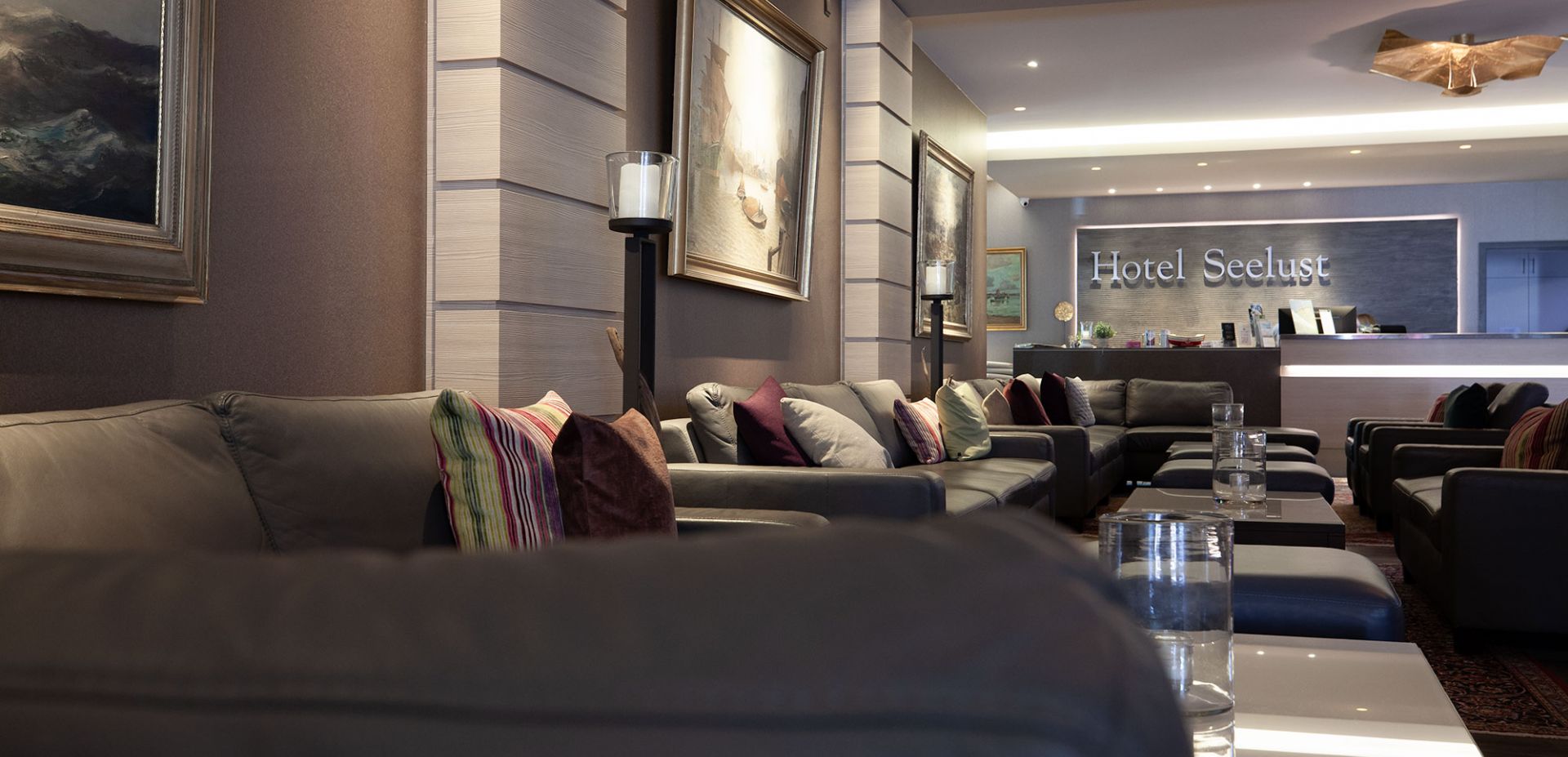 The lobby of the Hotel Seelust features gray sofas, colorful cushions, and glass-topped tables. Paintings hang on the walls, and the reception desk with a Hotel Seelust sign can be seen in the background.