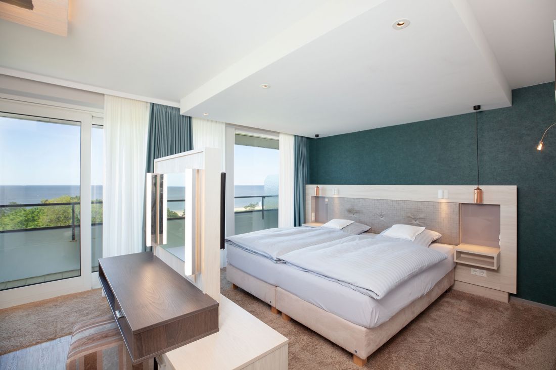 Modernes Zwei-Raum-Suite mit zwei Einzelbetten, einem großen Fenster mit durchsichtigen Vorhängen, einem Balkon, einem Schreibtisch mit Spiegel und Blick auf die Nordsee..