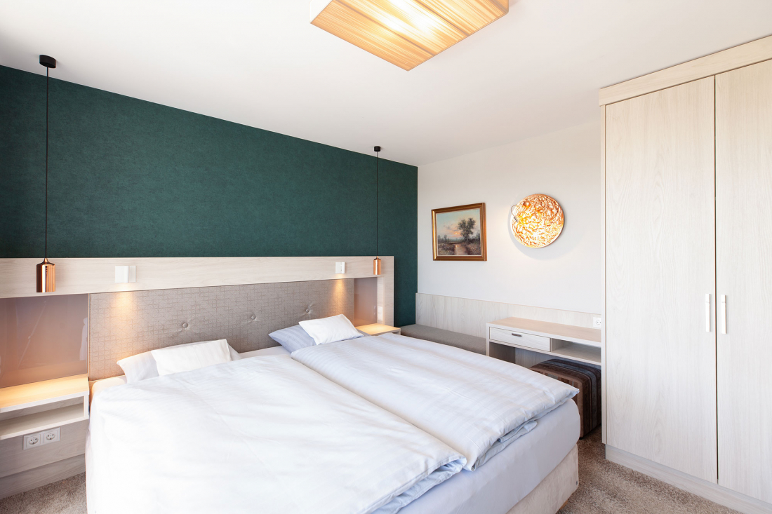 Schlafzimmer mit Doppelbett, einer blaugrünen Akzentwand, Pendelleuchten, einem Kleiderschrank, einem Schreibtisch und Wandkunst über einer Bank. Weißes Bettzeug und helle Holzmöbel sorgen für einen hellen, klaren Look.