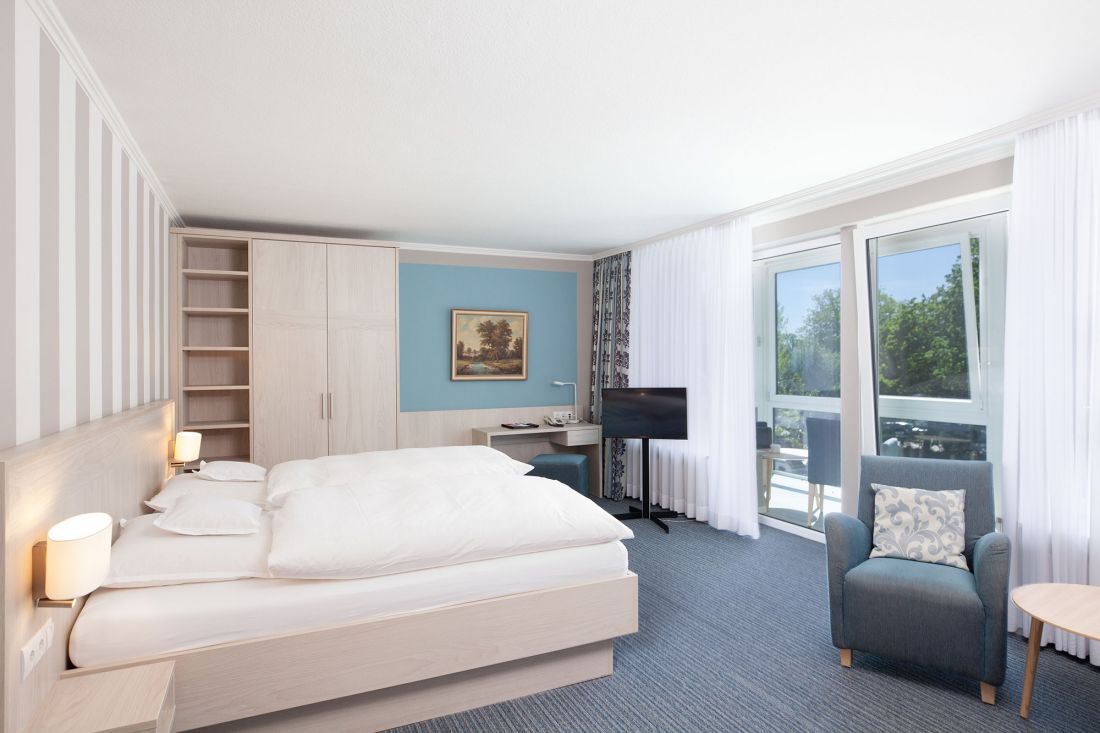 Ein helles Hotelzimmer mit einem Doppelbett, Sessel, Schreibtisch, Fernseher, großem Kleiderschrank und einem Fenster mit durchsichtigen Vorhängen mit Blick ins Grüne. Die Einrichtung ist hell mit blauen und weißen Tönen und einem Gemälde an der Wand.