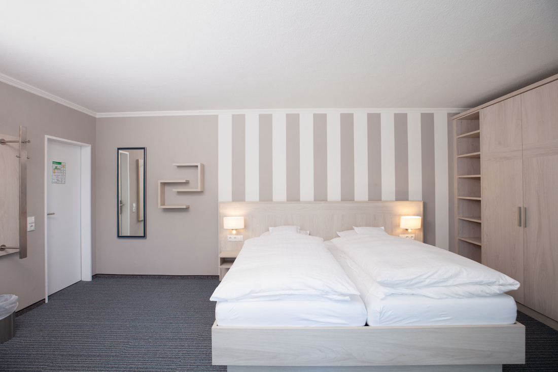 A modern hotel room with a double bed, white bed linen, two bedside lamps, open shelves, a wall-mounted mirror and a striped accent wall behind the bed. The design is minimalist with neutral colours.