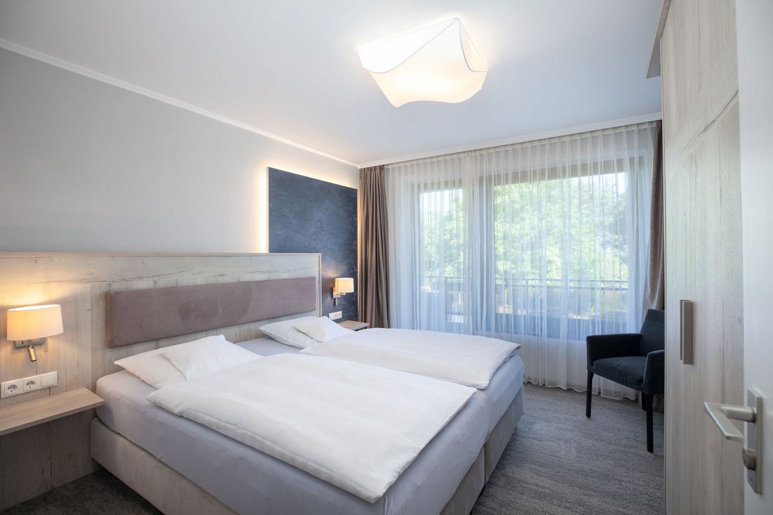 Schlafzimmer eines Dreiraum-Appartements mit Doppelbett, hellen Holzmöbeln, Nachttischlampen, einer dunklen Akzentwand, einem großen Fenster mit durchsichtigen Vorhängen und einem schwarzen Stuhl am Fenster.