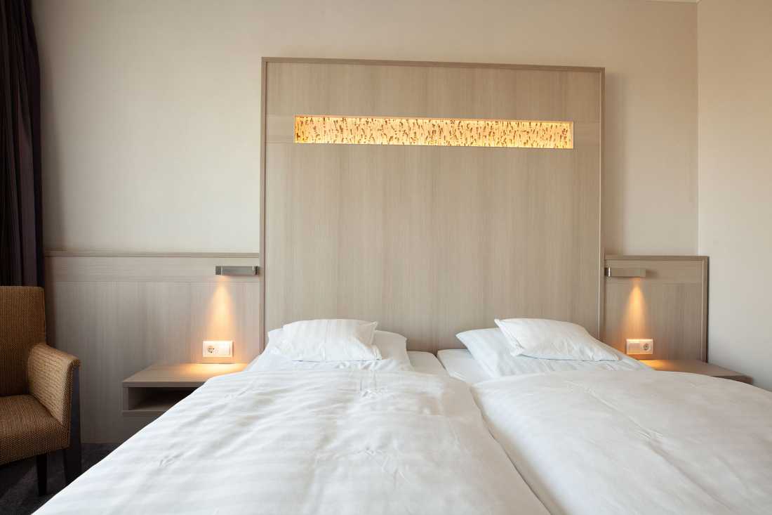 A double room with two single mattresses in a double bed, each with white bed linen and pillows. The headboard has built-in lamps and wood panelling. A brown armchair is partially visible on the left-hand side.