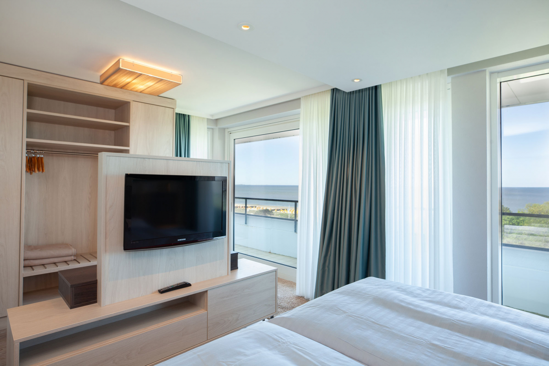 Schlafzimmer der Zwei-Raum-Suite mit Doppelbett, einem Fernseher auf einem hellen Holzständer, einem offenen Kleiderschrank und großen Fenstern mit durchsichtigen und dunklen Vorhängen, die einen Blick auf das Meer und einen Balkon nach draußen bieten.
