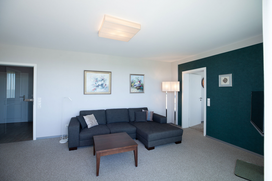 Modernes Wohnzimmer mit einem dunkelgrauen Sektionssofa, einem Couchtisch aus Holz, einer Stehlampe, einer türkisfarbenen Akzentwand und zwei abstrakten Kunstwerken, die über dem Sofa hängen. Im Hintergrund sind eine Tür und ein Flur zu sehen.
