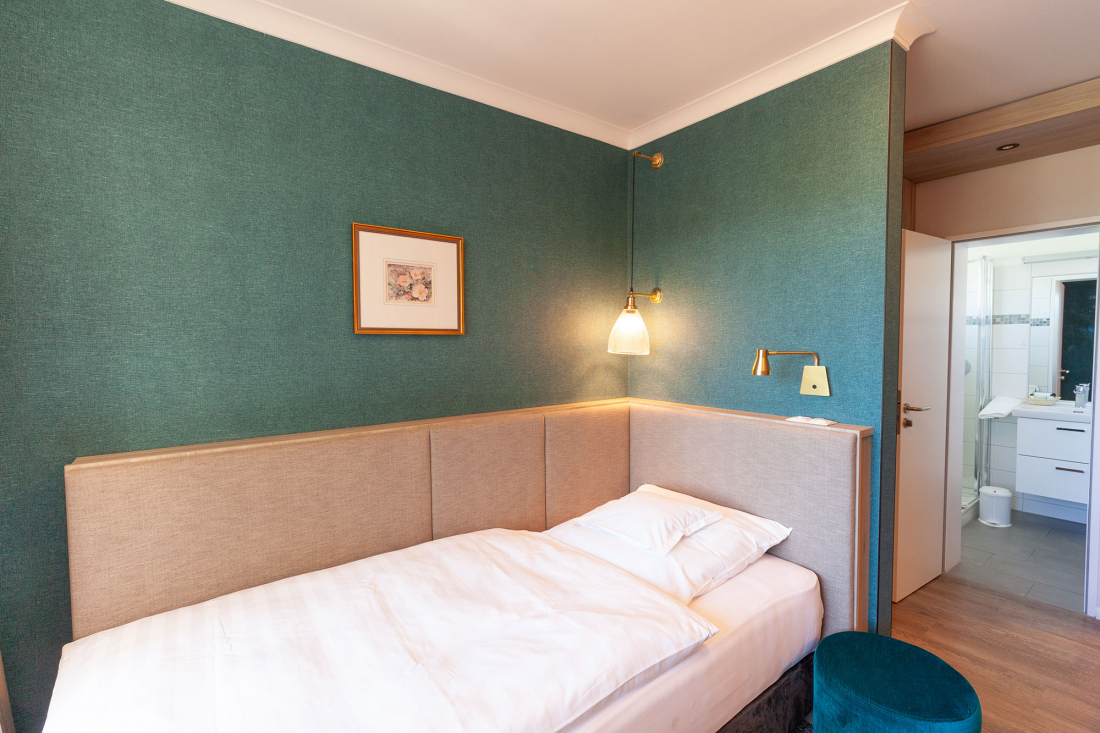 A small, modern hotel room with teal-coloured walls, a single bed with white bed linen, a wall lamp, framed artwork and an open door leading into a bright bathroom.