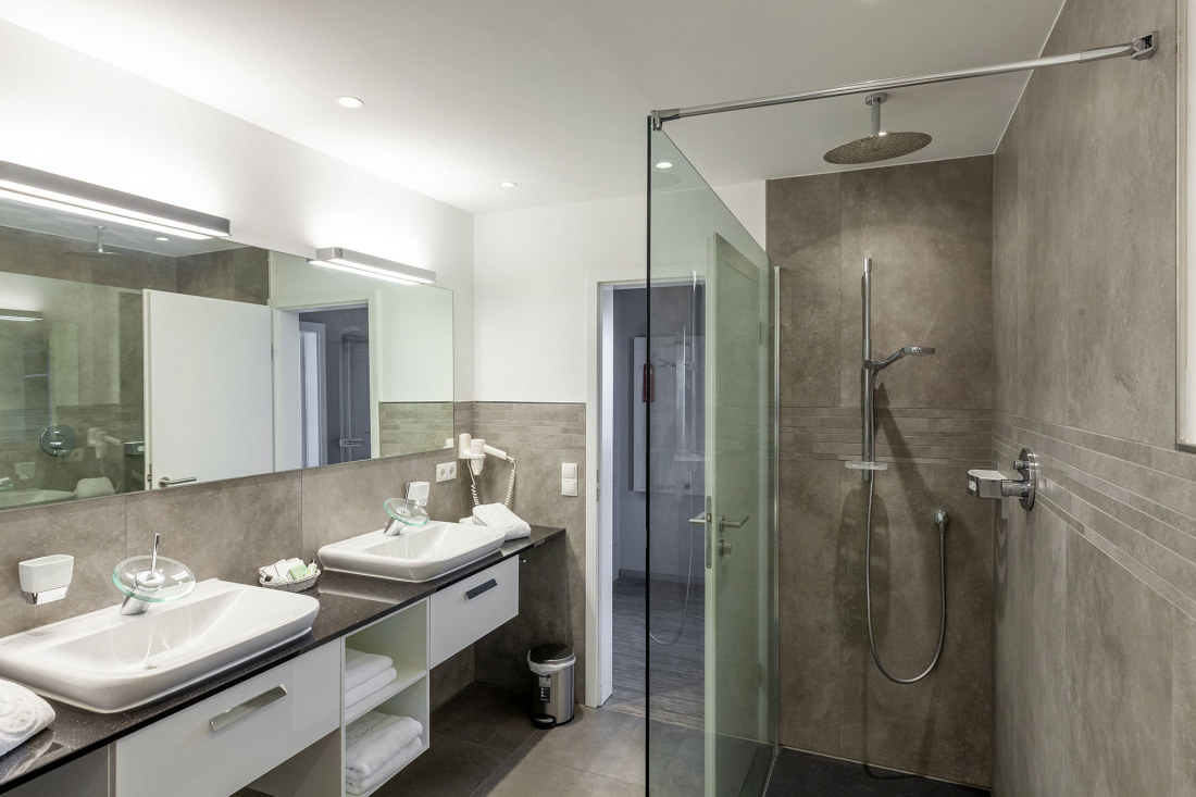 Modernes Badezimmer mit einem großen Spiegel, zwei Waschbecken mit Toilettenartikeln, weißen Schränken mit Handtüchern, einem an der Wand befestigten Haarföhn, einer gläsernen, begehbaren Dusche mit Regenduschkopf und neutralen grauen Fliesen an Wänden.