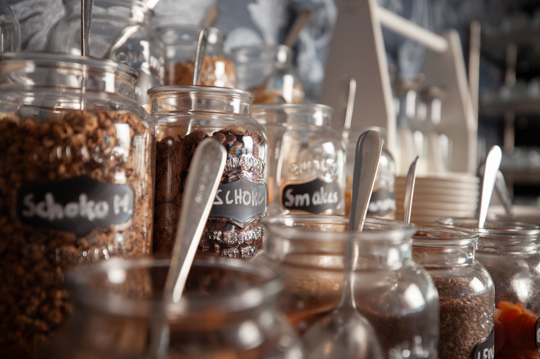 Eine Reihe von Gläsern mit verschiedenen Cerealien und Toppings, jedes mit einem Löffel und einem kleinen Kreidetafel-Etikett, die auf einer Frühstückstheke oder einem Buffet stehen.