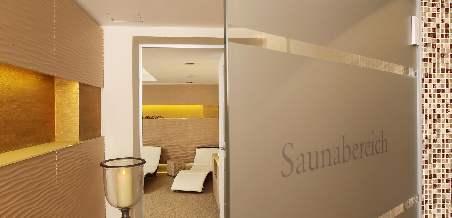 Moderner Wellnessbereich mit beigefarbenen, strukturierten Wänden, warmer Beleuchtung und weißen Liegestühlen. Eine Glastür mit der Aufschrift "Saunabereich" ist teilweise geöffnet und lädt zum Eintritt in den Entspannungsraum ein.