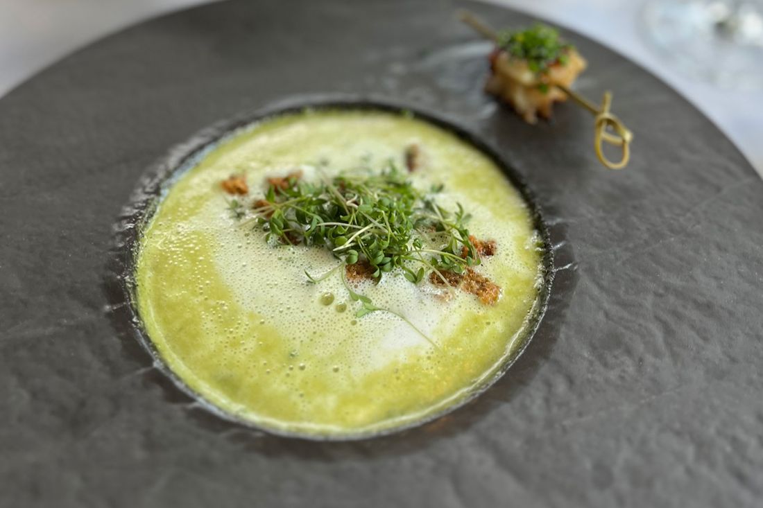 A bowl of green cream soup garnished with fresh sprouts and croutons, served on a dark plate with a skewer of grilled food and microgreens on the side.