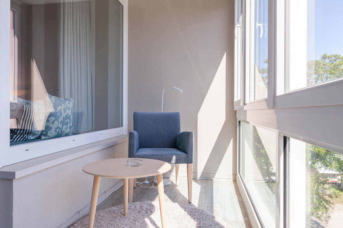 A bright, modern balcony with large windows, a grey armchair, a small round wooden table with an ashtray, a floor lamp and a soft beige carpet. The sunlight streams in and creates a cosy atmosphere.