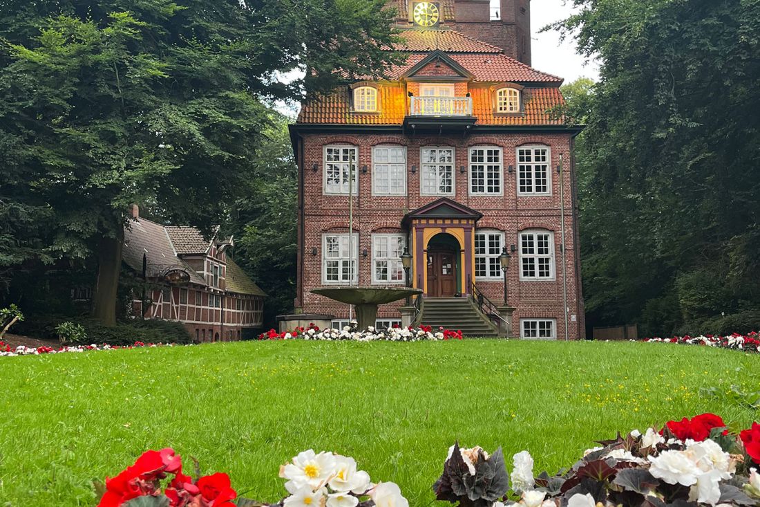 Schloss Ritzebüttel, ein Backsteingebäude mit weiß umrandeten Fenstern steht am Ende eines grasbewachsenen Rasens, umgeben von Bäumen und rot und weiß blühenden Blumenbeeten. Vor dem Eingang befindet sich ein kleiner Springbrunnen.