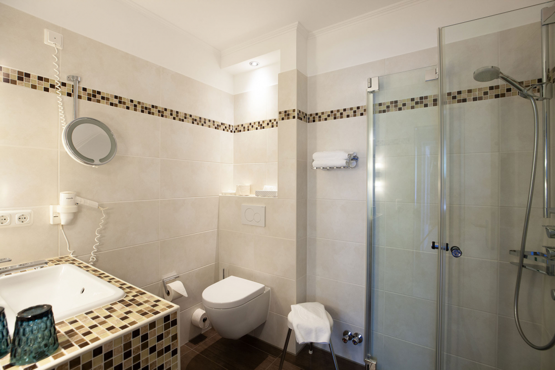 Modernes Badezimmer mit beigen Fliesen, einer an der Wand befestigten Toilette, einer Glasduschkabine, einem Waschbecken mit Mosaikarbeitsplatte, einem runden Spiegel, einem montierten Haartrockner, Handtüchern und einer sanften Deckenbeleuchtung.