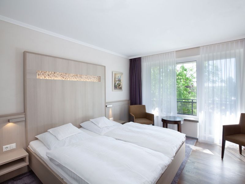 A bright, modern hotel room with two single beds pushed together, white bedding, a wooden headboard, two brown chairs at a round table, a large window with sheer curtains and a painting on the wall.