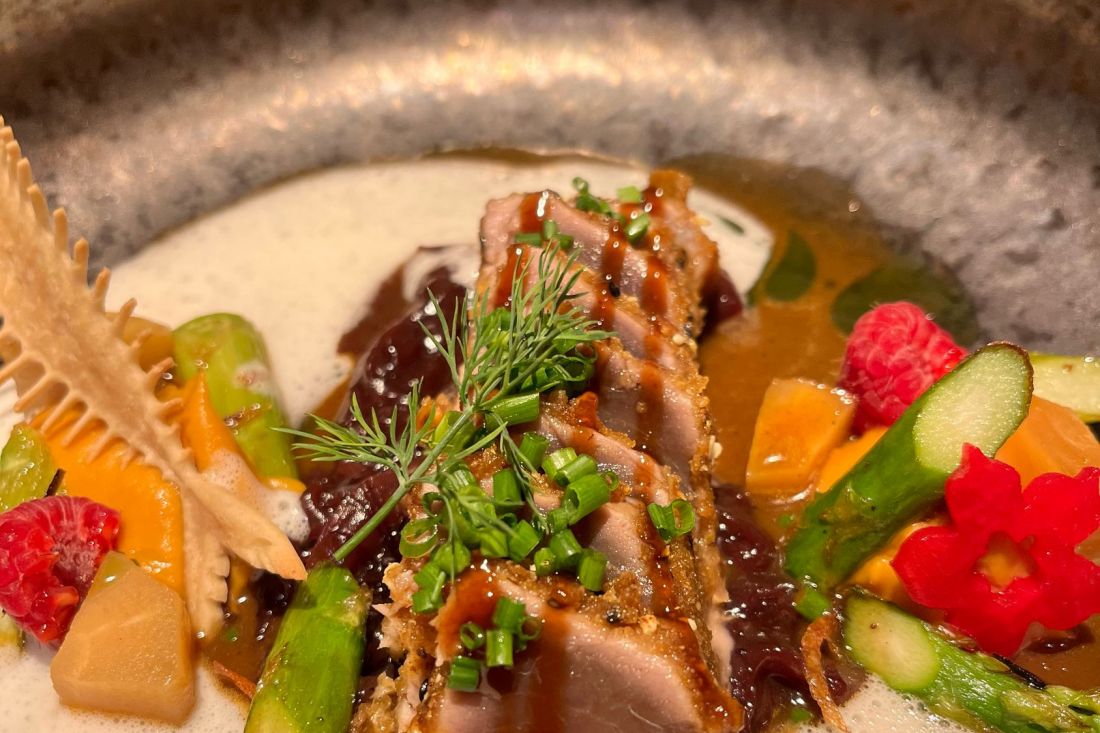 Close-up of a gourmet dish with seared tuna slices and herbs, surrounded by asparagus, carrots, raspberries, a creamy sauce, and a decorative crispy piece in a stoneware bowl.