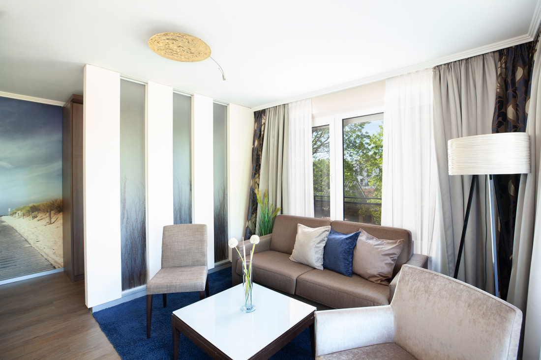 Helles, modernes Wohnzimmer eines Zweiraum-Appartements mit einem Sofa, zwei Sesseln, einem Couchtisch, einer Stehlampe und großen Fenstern mit transparenten Vorhängen. Dekorative Paneele und eine Wandmalerei mit einer Strandszene verschönern den Raum.