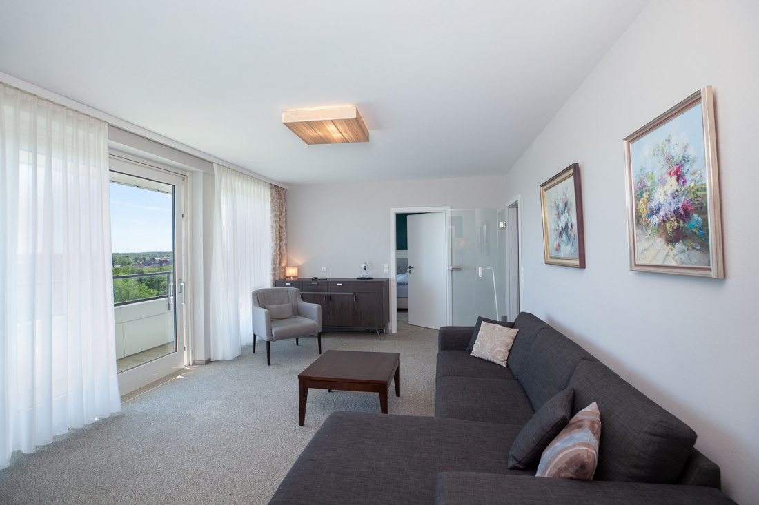 Ein helles, modernes Wohnzimmer mit einem grauen Sofa, einem hölzernen Couchtisch, einem Schreibtisch und einem Stuhl, großen Fenstern mit durchsichtigen Vorhängen und zwei bunten Blumenbildern an weißen Wänden. Eine Tür führt in ein anderes Zimmer.