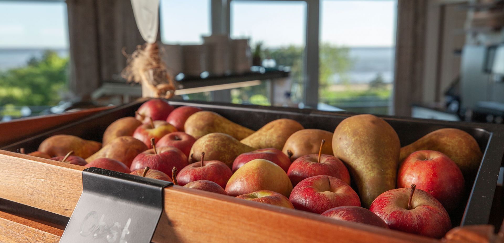 Apples in a dark wooden box with a black sign reading "Fruit." In the background is a large window overlooking the North Sea.